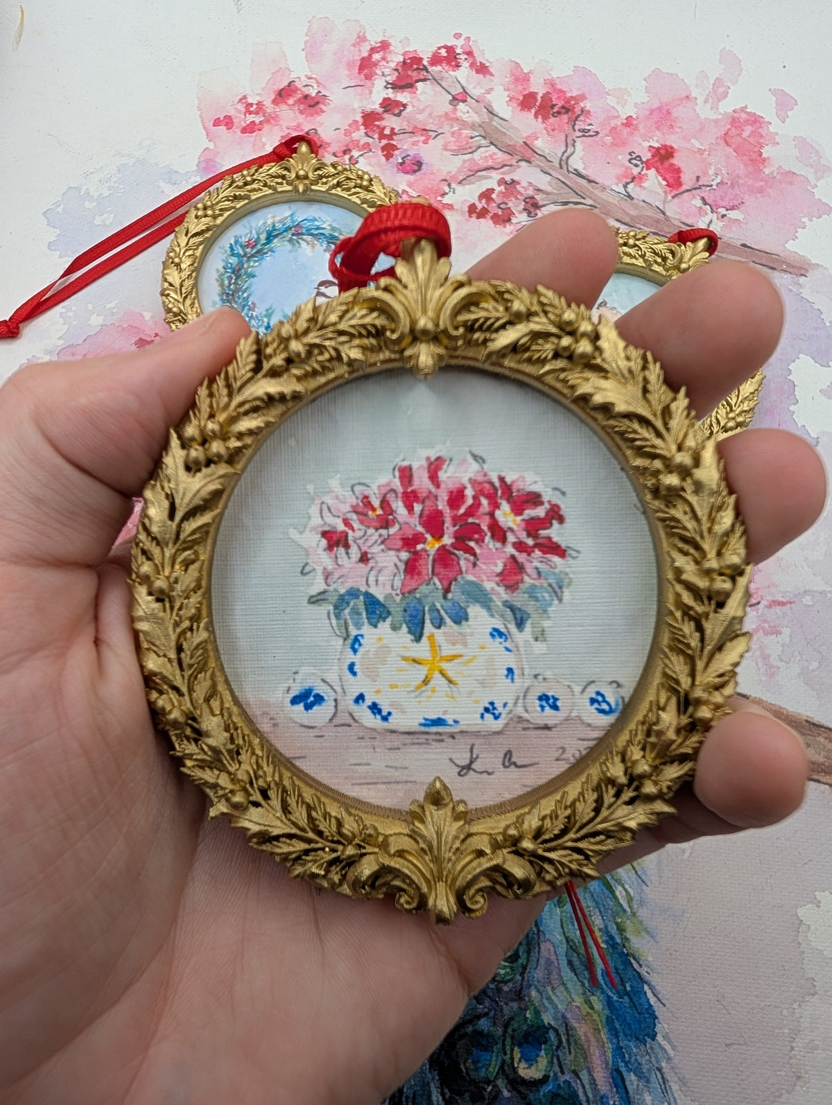 Poinsettias in a Blue and White Pot watercolor ornament in a gold wreath frame, held in hand